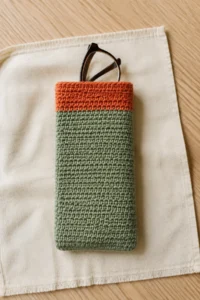 Sage crochet glasses sleeve with a terracotta top band and brown reading glasses on ivory linen.